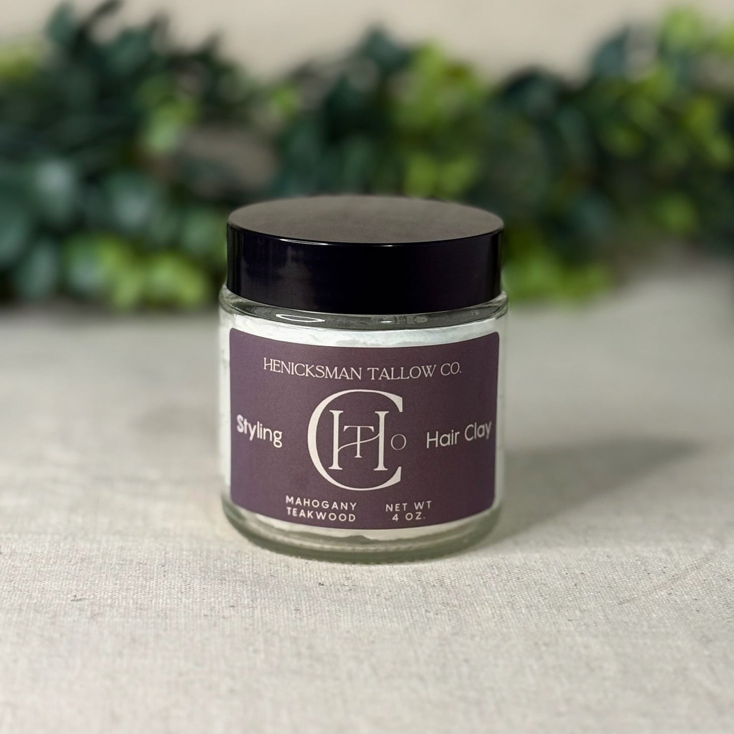 Jar of Henricksman Tallow Co. hair clay on a textured surface with greenery in the background
