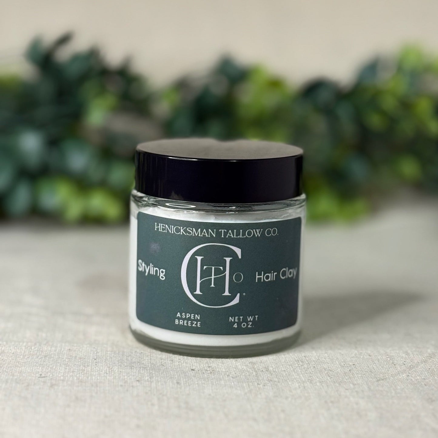 Jar of Henricksman Tallow Co. hair clay on a neutral background with greenery.
