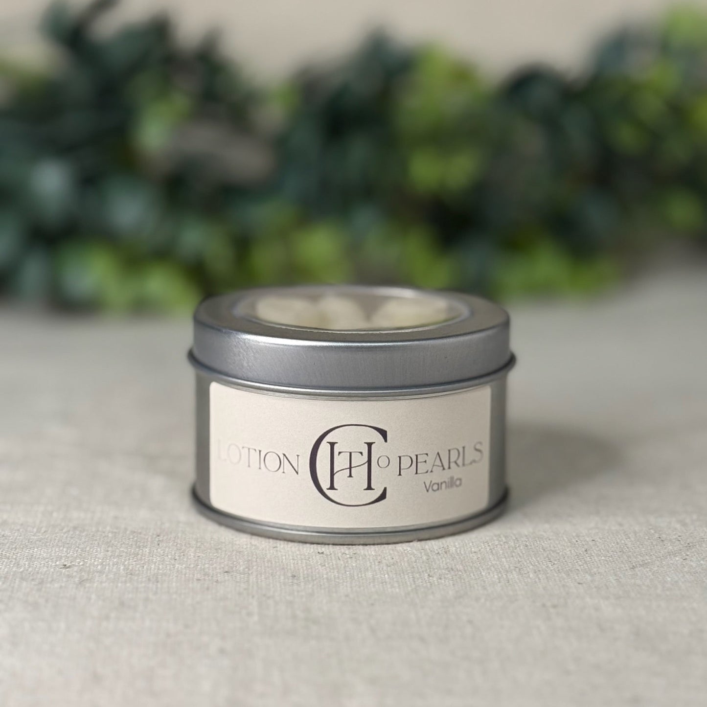 Candle tin labeled 'Pearls Vanilla' on a textured surface with blurred greenery in the background