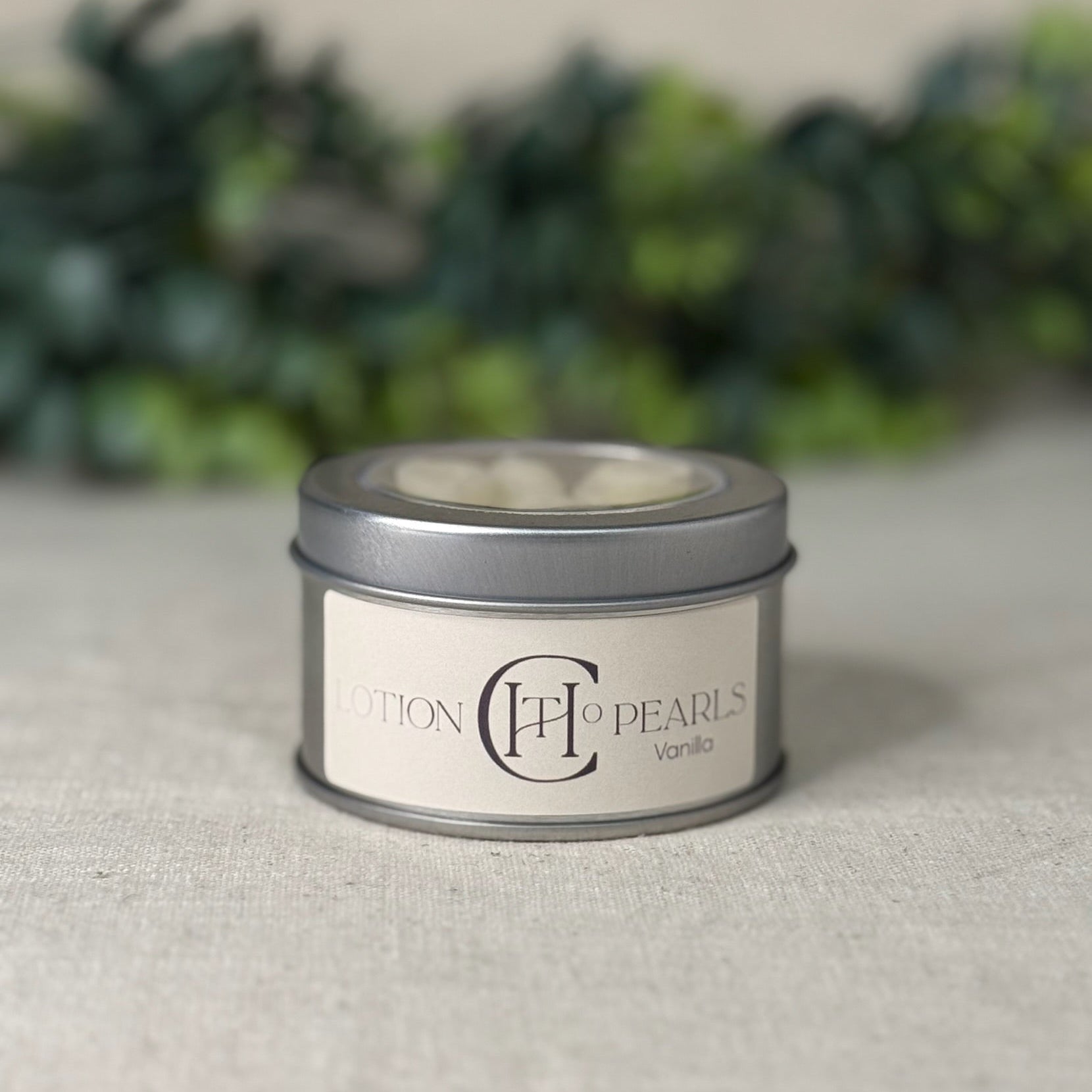 Candle tin labeled 'Pearls Vanilla' on a textured surface with blurred greenery in the background