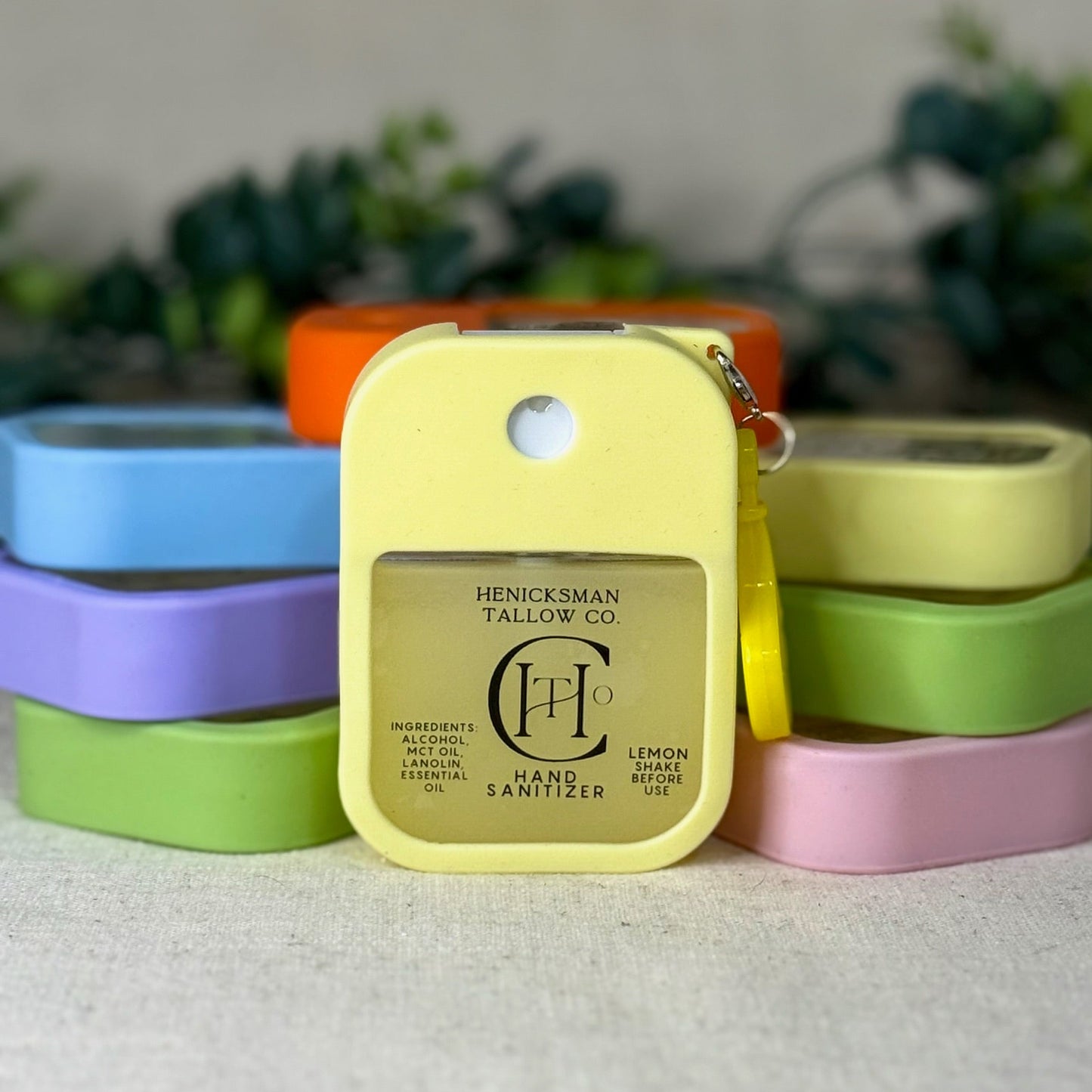 Yellow hand sanitizer bottle with 'Henicksman Tallow Co' branding against a blurred background of colorful containers.