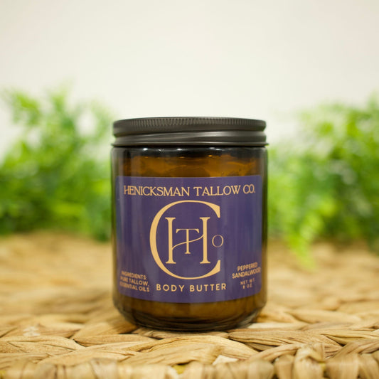 An amber glass jar of Henicksman Tallow Co. Body Butter with a dark purple label and black lid, set on a woven straw mat with a soft-focus green background.