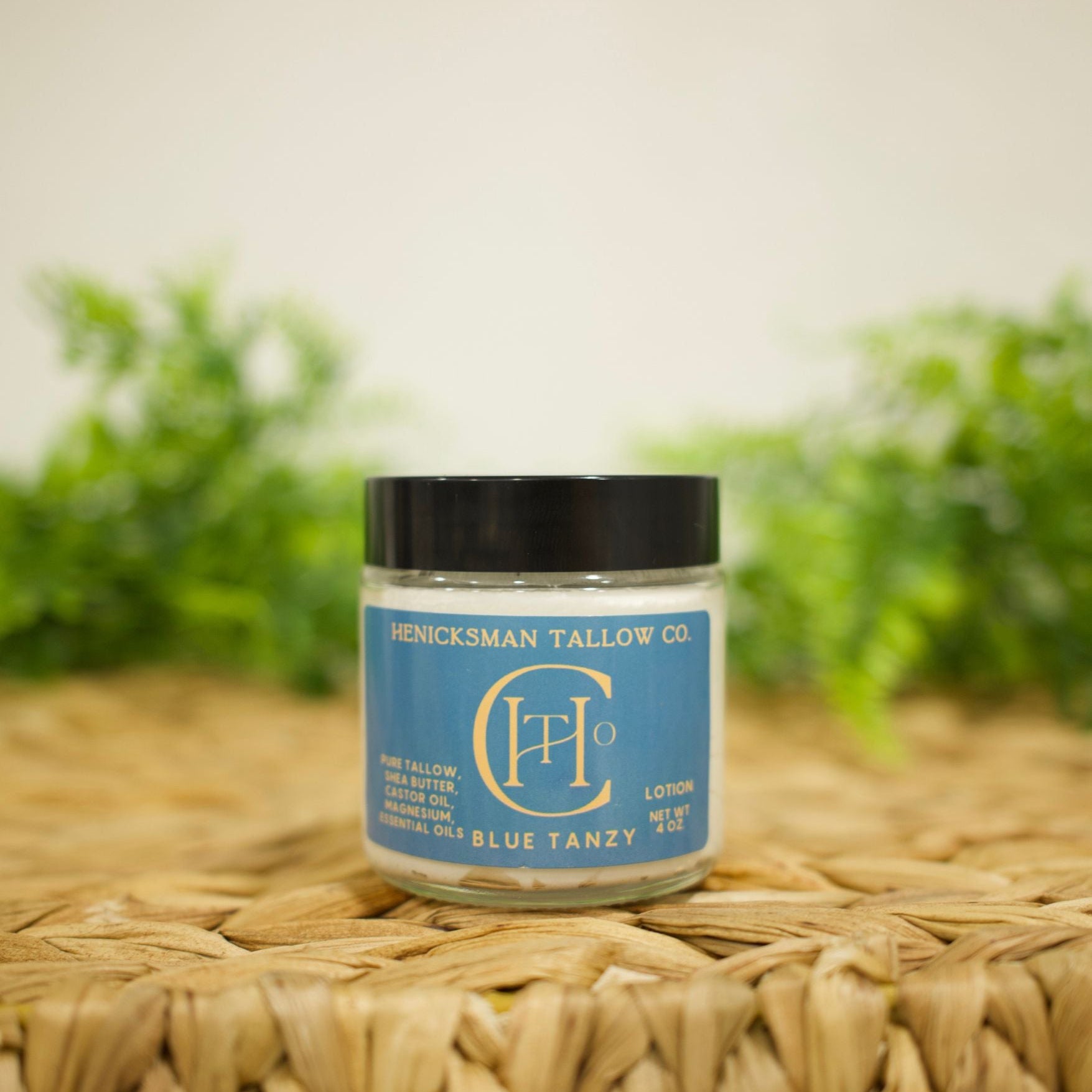 A jar of Henck'sman Tallow Co. 'Blue Tanzy' pure tanzy wax melt, featuring a blue label and black lid, displayed on a woven straw mat with a soft-focus green background.
