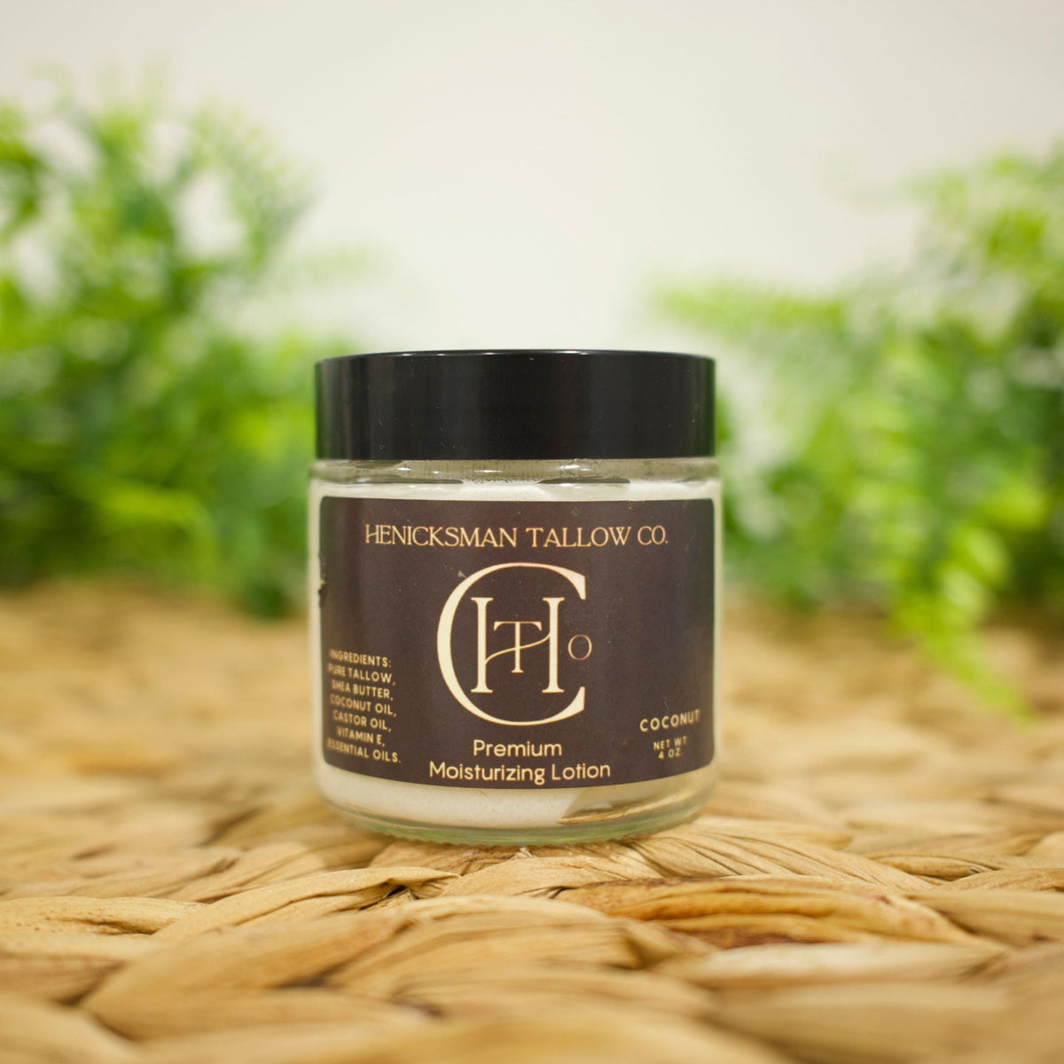 A jar of Henicksman Tallow Co. Premium Moisturizing Lotion in coconut scent, placed on a woven straw mat with a soft-focus green background.