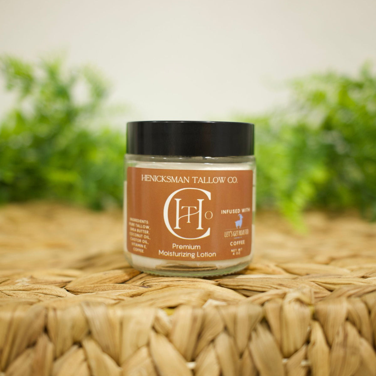 A jar of Henckelsman Tallow Co. nerased wax moisturizing lotion sits on a woven straw mat with a blurred green plant background.