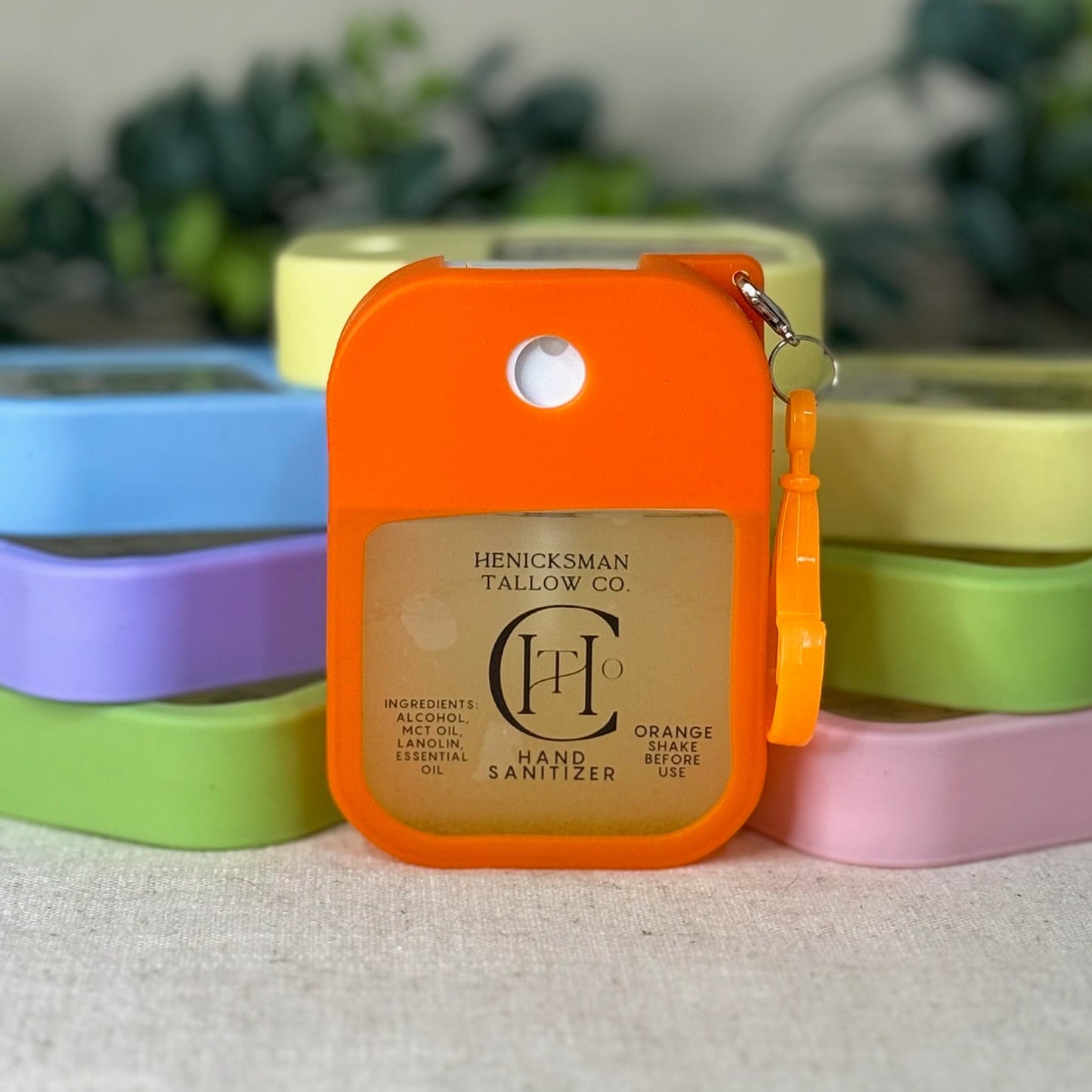 Orange hand sanitizer with 'Henicksman Tallow Co.' branding on a white surface with colorful containers in the background.
