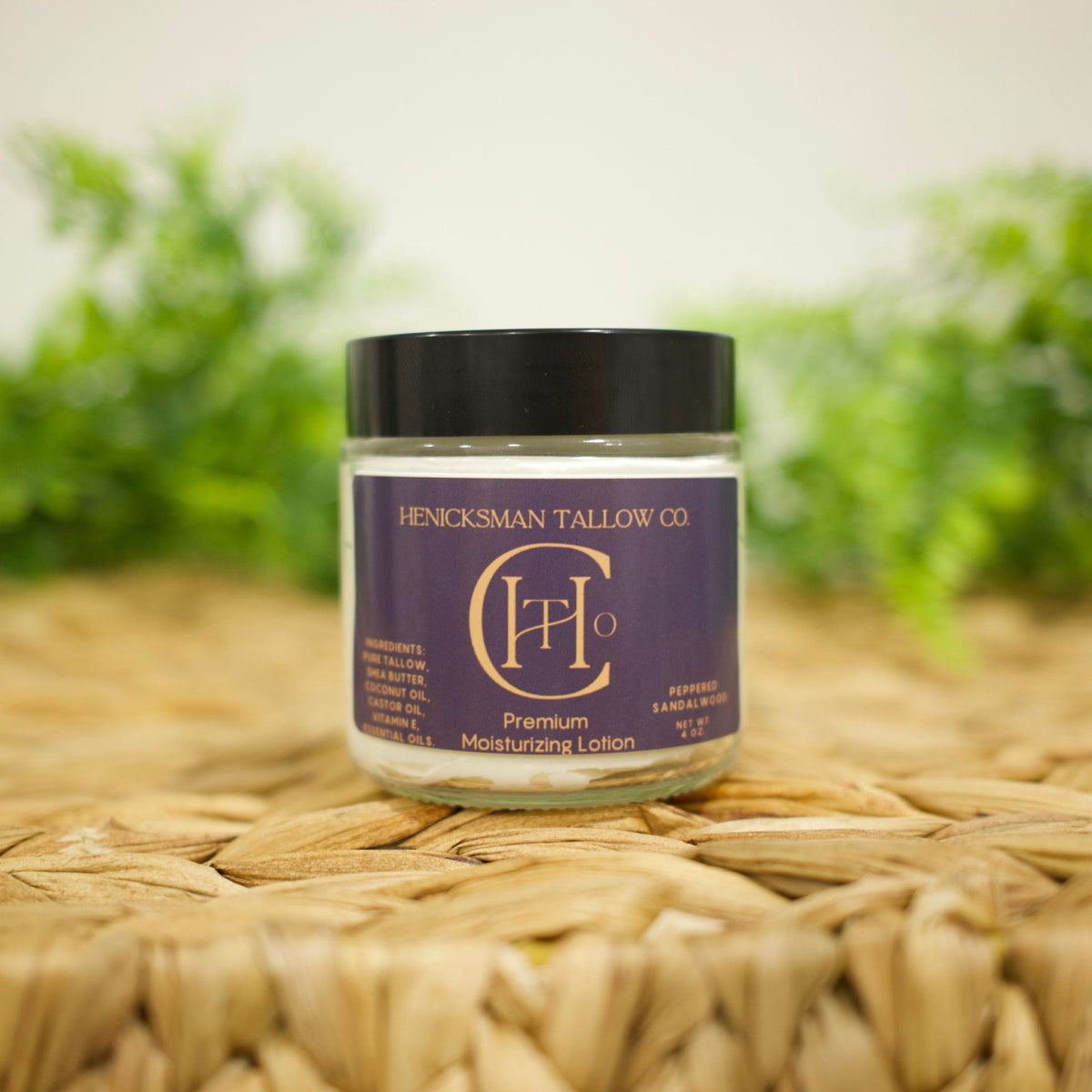 A jar of Henicksman Tallow Co. Premium Moisturizing Lotion with a black lid and a purple label, resting on a woven straw mat with a soft-focus green background.