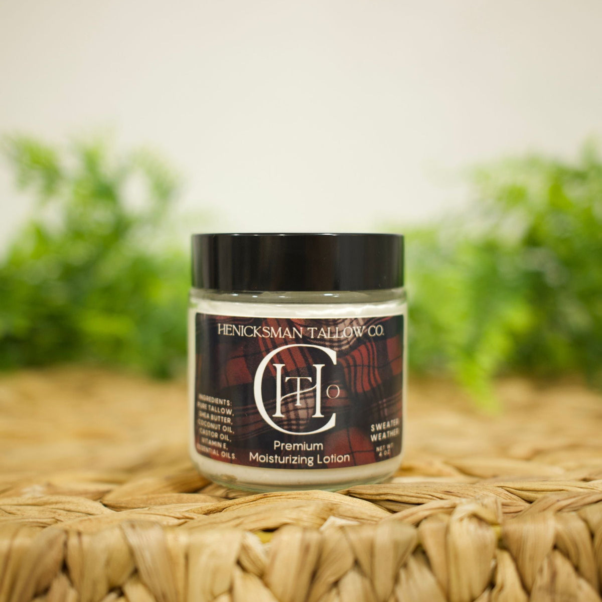 A jar of Henckelsman Tallboy Co CH20 Premium Moisturizing Lotion with a black lid and dark label, resting on a woven straw mat with a blurred green background.