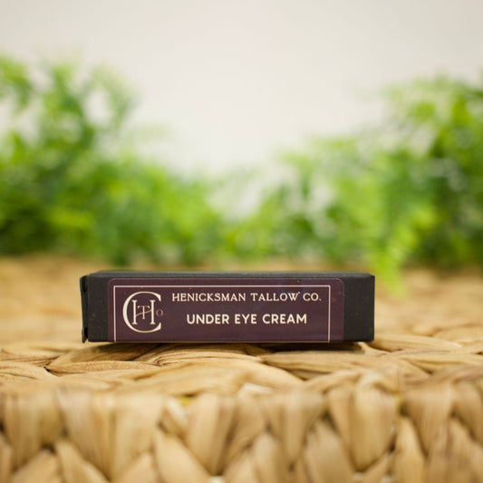 Henicksman Tallow Co. Under Eye Cream in a dark rectangular box, resting on a woven straw mat with a blurred green and white background.
