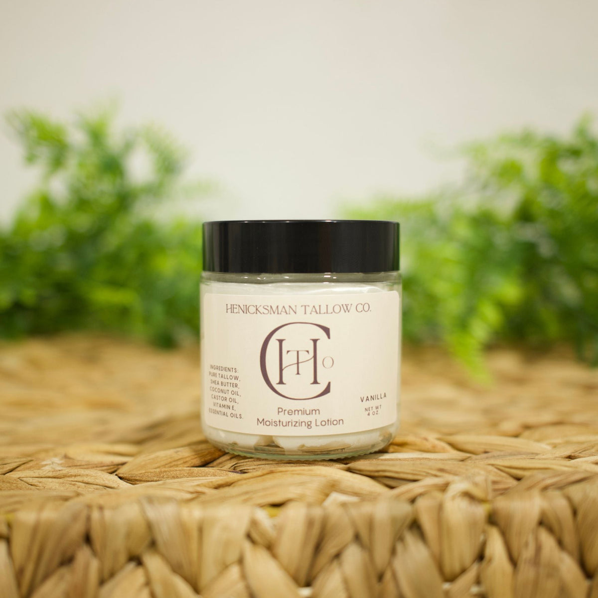 A jar of Henckelsman Tallow Co. Vanilla Premium Moisturizing Cotion sits on a woven straw mat with a soft-focus background of green foliage and a light wall.