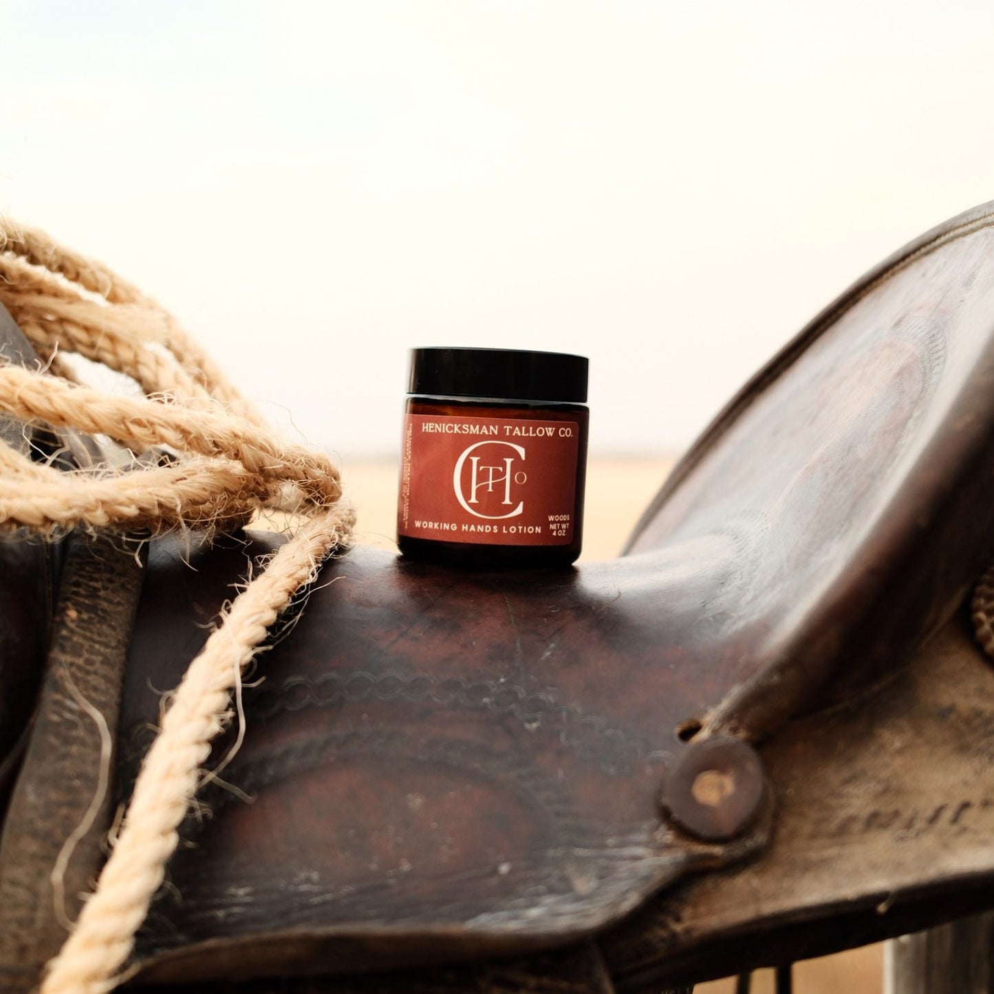 A small jar of Shockwave Tallow Co. hair product sits on the leather strap of a brown western saddle with rope in the foreground, set against a blurred outdoor background.